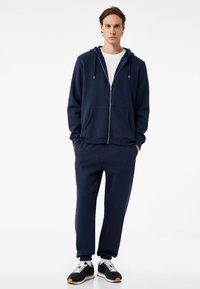 Marineblå zip-up hoodie med to forlommer, parret med matchende joggers. Sættet har en blød tekstur og er lavet af et tykt stof.