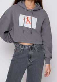 Kobieta w szarej, skróconej bluzie Calvin Klein i szarych, wysokich dżinsach, stojąca z jedną ręką na biodrze na tle jednolitego koloru.