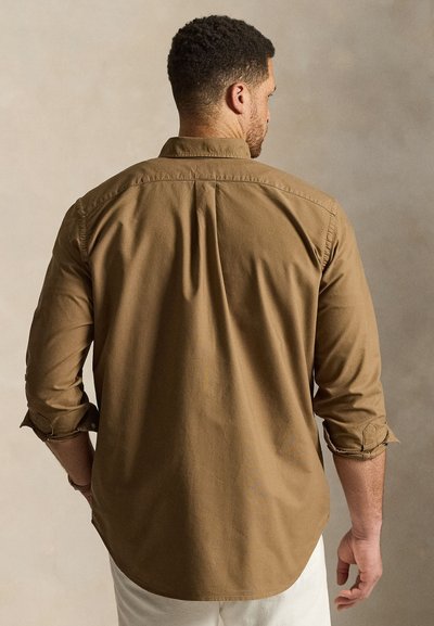 Mężczyzna z podwiniętymi rękawami, ubrany w brązową koszulę z długim rękawem i jasne spodnie, stojący na tle jednolitego neutralnego tła, zwrócony tyłem.