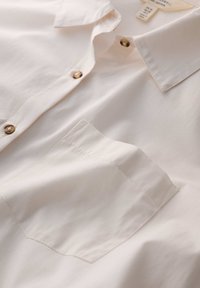 Jasno beżowa koszula zapinana na guziki, wykonana z bawełny, z kołnierzykiem w szpic, złotymi guzikami i jedną kieszenią na piersi o prostokątnym kształcie.