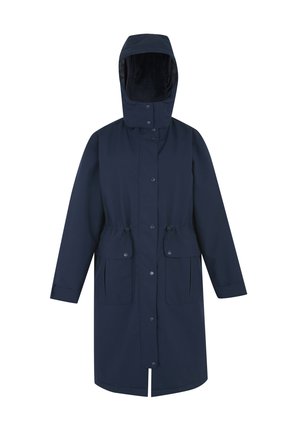 Kobieta w beżowej dzianinowej czapce, granatowym długim płaszczu, białym swetrze, niebieskich dżinsach i białych trampkach z czarnymi i beżowymi akcentami.