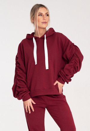 Bordowa bluza z kapturem, z białymi sznurkami, w oversize'owym fasonie, z kieszeniami i marszczonymi rękawami dla tekstury. Noszona z dopasowanymi spodniami.