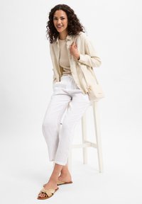 Light beige button-up shirt over a beige top, white linen pants, and light brown sandals with a knot detail. Small cream handbag with a strap.