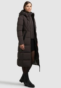 Langer, dunkelbrauner Puffer-Mantel mit Kapuze, ausgestattet mit horizontalen Nähten, Reißverschlusstaschen und einer glatten Textur. Kombiniert mit schwarzen Ankle Boots.