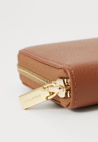 Portefeuille en cuir texturé marron avec fermeture éclair dorée et quincaillerie, comportant une étiquette de marque visible près du tirette de la fermeture. Forme rectangulaire compacte.