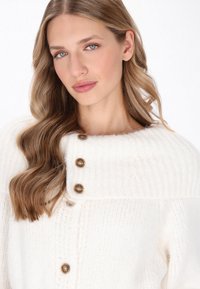 Crème-kleurige gebreide cardigan met een hoge kraag, drie bruine knopen aan de voorkant en een zachte, gestructureerde stof.