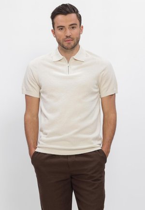 Man met kort donker haar, die een beige korte mouw polo met rits draagt en bruine broek, staand met handen in de zakken.