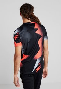 Man med dreadlocks som bär en svart träningsskjorta med ett abstrakt röd- och vitblixtrande mönster, sedd bakifrån.