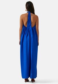Robe maxi bleue à halter avec un dos nu, dotée d'un nœud au cou et d'un tissu léger et fluide. Associée à des escarpins rouges.
