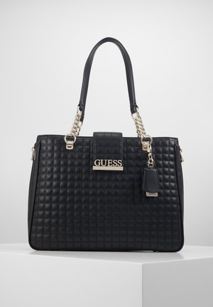 Sac fourre-tout en cuir matelassé noir avec des poignées en chaîne dorée et une plaque logo "GUESS" à l'avant, posé sur un socle blanc.