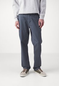 Pantalon en coton bleu marine à coupe décontractée, avec une taille plate, des poches latérales et une texture lisse, associé à des baskets grises et olive.