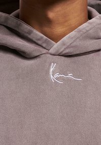 Hanorac gri deschis cu un finisaj texturat, având un logo brodat alb pe piept și o glugă cu siret.