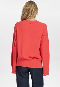 Koralen pleteni pulover s sproščenim krojem, z rebrastimi manšetami in robom ter majhnim gumbom na hrbtni strani. V kombinaciji s temnimi jeans hlačami.