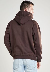 Felpa con cappuccio marrone con finitura texture, presenta un grande cappuccio, maniche raglan e orlo a coste. La parte posteriore è semplice.