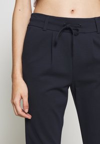 Marinblå byxor med dragsko i midjan, veckad frontdesign och mjukt, texturerat tyg. Närbild som visar detaljer i passform och material.