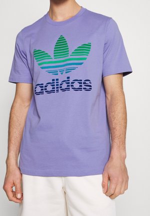 Mann trägt ein helllila Adidas-T-Shirt mit grün-blau gestreiftem Trefoil-Logo und weißen Shorts vor einfarbigem Hintergrund.