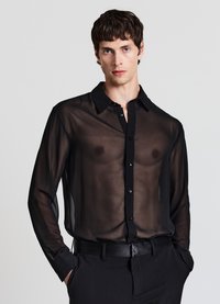 Schwarze, transparente Bluse mit klassischem Kragen, langen Ärmeln und dezenten Knopfdetails, getragen mit einem schwarzen Gürtel und eleganter Hose.