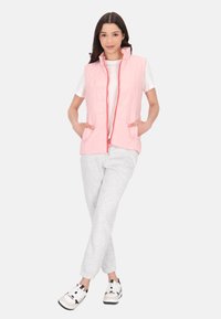Gilet piumino rosa chiaro con colletto alto, chiusura con zip e due tasche laterali, indossato sopra una camicia bianca e pantaloni sportivi grigi.
