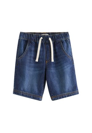 Ung dreng smiler, iført en beige t-shirt med et zebra motiv, denim shorts med snore, gule sneakers og lyse sokker.