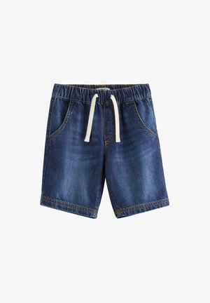 Ung dreng smiler, iført en beige t-shirt med et zebra motiv, denim shorts med snore, gule sneakers og lyse sokker.