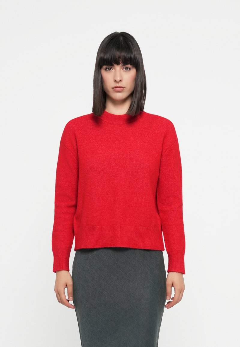 Red woolen sweater with a ribbed neckline and cuffs, featuring a straight cut and a slightly cropped length. Worn with a dark pencil skirt.