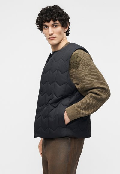 Νεαρός άνδρας με σγουρά μαλλιά που φοράει μαύρο γιλέκο με ραφές πάνω από πράσινη ελιά υφασμάτινη μπλούζα με υφασμάτινο λουλουδάτο μοτίβο, στημένο με το ένα χέρι στην τσέπη.