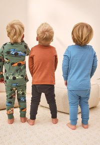 Trois enfants sont debout, dos à la caméra, portant des pyjamas colorés : vert avec des motifs de voitures, rouille et bleu clair. Tous ont les cheveux clairs.