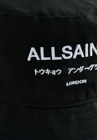 Musta puuvillainen hattu, jossa on valkoista brodeerattua tekstiä "ALLSAINTS" ja muita merkkejä sekä näkyviä ompelut yksityiskohtia.