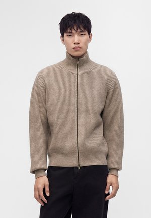 Jeune homme portant un pull côtelé beige avec fermeture éclair et un pantalon noir, debout devant un fond clair uni.