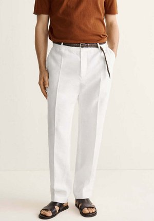 Hombre con pantalones blancos plisados, cinturón trenzado marrón, camisa marrón de manga corta y sandalias negras con punta abierta, de pie con las manos en los bolsillos.