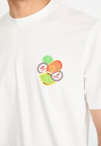 White cotton t-shirt featuring colorful, circular graphic logos in green, red, orange, and brown on the upper left chest.