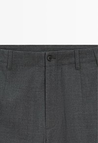 Primo piano di pantaloni eleganti grigio scuro che mostrano la vita con bottone, passanti per cintura e zona della cerniera frontale.