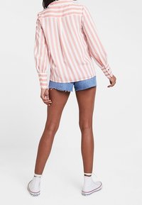 Camisetta a maniche lunghe a righe rosa e bianche con polsini abbottonati, abbinata a pantaloni corti in denim blu e sneakers bianche. Vista posteriore che mostra la vestibilità e le lunghezze.