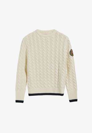 Pull crème en maille torsadée avec poignets et ourlet côtelés, bordure noire, et écusson brodé sur la manche gauche.