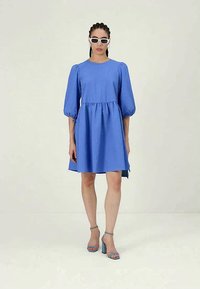 Egy nő textúrált, térdőig érő kék ruhát visel, puffos ujjakkal, fehér napszemüveget és kék blokksarkú szandált, egy egyszínű háttér előtt.