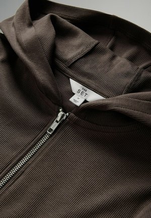 Braune gerippte Kapuzenjacke mit silbernem Reißverschluss, einem Etikett mit der Aufschrift "THE SET" und einem strukturierten Stoff. Die Kapuzenjacke hat einen lässigen Kragen.
