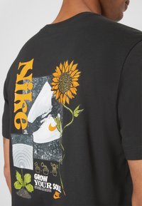Svart t-shirt med orange "Nike" text, vit sneakers grafik, solros, växtillustrationer och "Grow Your Sole" text på baksidan.