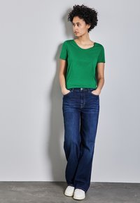Zelené tričko s krátkým rukávem spárované s volnými tmavě modrými džíny a bílými teniskami, s kulatým výstřihem a pohodlným střihem.