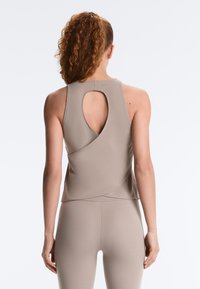 Vrouw met krullend haar in een hoge paardenstaart, gekleed in een beige mouwloos shirt met een sleutelgat aan de achterkant en bijpassende leggings, staand tegen een witte achtergrond.