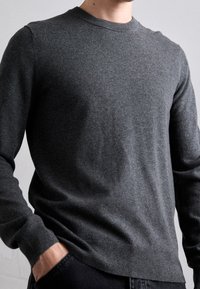 Grå stickad tröja med långa ärmar och rund halsringning. Mjuk textur, figurnära design, ribbade ärmslut och nederkant. Bärs över mörka byxor.