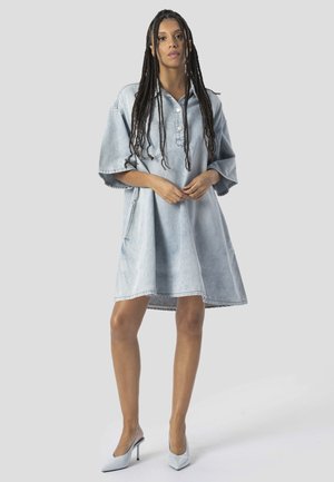 Mujer con trenzas largas que lleva un vestido vaquero holgado de color azul claro y zapatos blancos de tacón puntiagudos, de pie sobre un fondo gris.