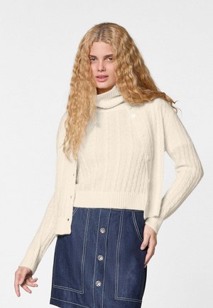 Vrouw met lang, krullend blond haar draagt een crème kabelgebreide coltrui en een bijpassend vest over een donkere denimrok met knopen.