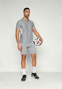 Hall mustas-valge jalgpall, seljas hall sportlik riietus, valged sokid ja mustad spordijalatsid, seistes ühtlase tausta ees.