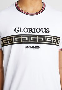 Valkoinen puuvillatee paita, jossa musta "GLORIOUS" -teksti ja kultainen geometrinen kuvio rinnan kohdalla, mukana raidallinen punainen ja musta kaulus.
