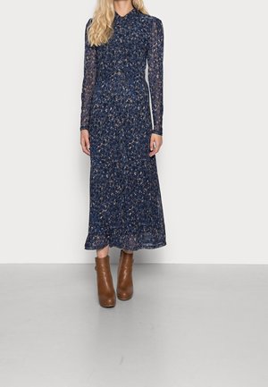 Femme portant une robe midi à manches longues, bleu foncé à motif floral, associée à des bottines marron, debout devant un fond clair uni.