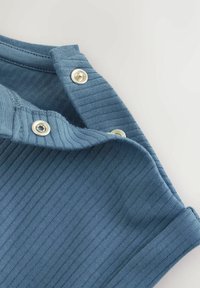 Blå ribbad tyg med metalliska tryckknappar på axeln. Materialet verkar mjukt och texturerat, vilket framhäver en avslappnad design.