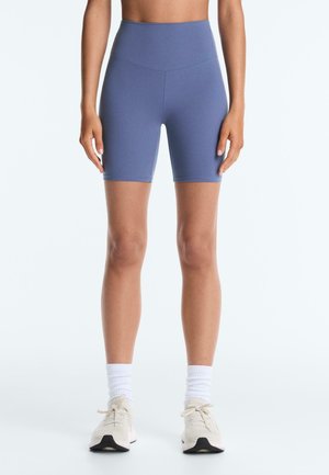 Femeie care poartă pantaloni scurți de biker albăstri cu talie înaltă, șosete albe și adidași bej, stând cu mâinile relaxate pe lângă corp.