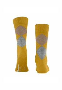Para skarpetek w musztardowym kolorze z wzorem argyle w brązowym i szarym, jedna skarpetka pokazuje spód z białym napisem marki.