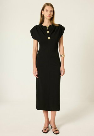Femme portant une robe midi noire avec des épaules structurées, des boutons décoratifs dorés, un bracelet en or, des boucles d’oreilles et des talons noirs à brides, debout sur fond blanc.
