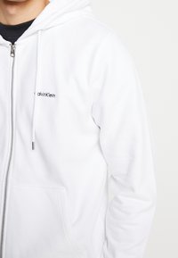 Man som bär en vit zip-up hoodie från Calvin Klein över en svart t-shirt, som visar överkroppen och en del av hans ansikte.